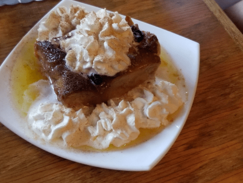 A slice of bread pudding topped with whipped cream and drizzled with sauce, served on a white plate.