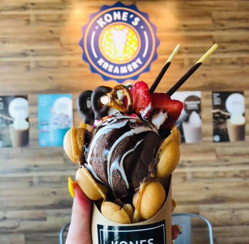 A hand holding a cone filled with chocolate ice cream, strawberries, and toppings, against a wooden wall with a logo.