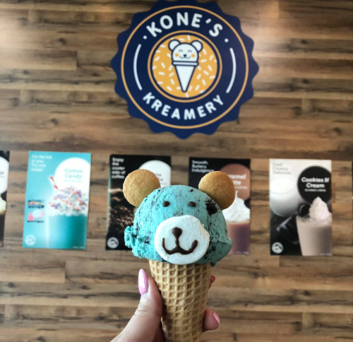 A hand holds a bear-shaped ice cream cone with blue and black ice cream, in front of a Kone's Kreamery sign.