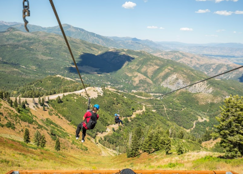 Two people ziplining over a mountainous landscape with green hills and blue skies in the background.
