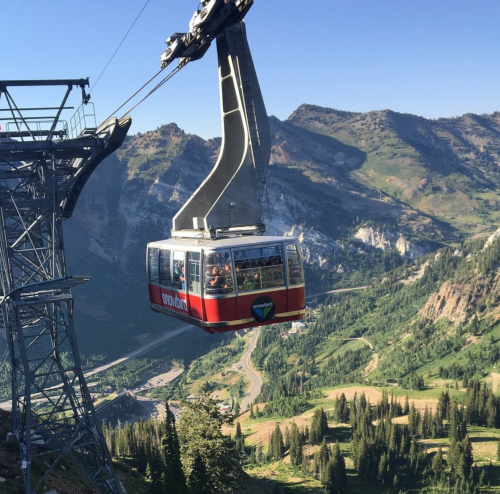 A gondola lifts over a mountainous landscape, surrounded by lush greenery and rocky peaks under a clear blue sky.