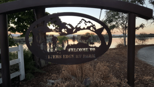 A metal sign reading "Welcome to Rivers Edge RV Park" with a scenic view of trees and water in the background.