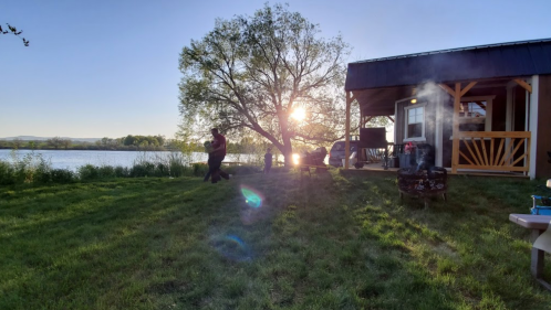 A serene lakeside scene at sunset, featuring a couple dancing near a tree and a cozy cabin with smoke rising from a fire.