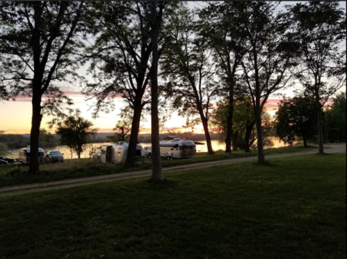 A serene sunset over a river, framed by trees, with campers visible in the background.