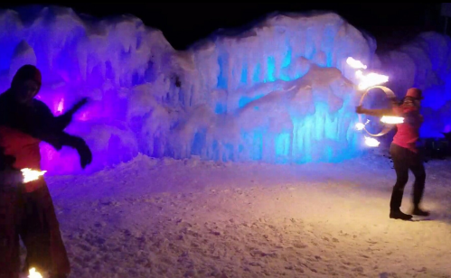 Two performers twirl fire props in front of a colorful ice wall, illuminated in blue and purple lights.