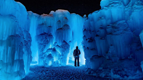 A person stands in a stunning ice cave illuminated by blue light, surrounded by towering ice formations.