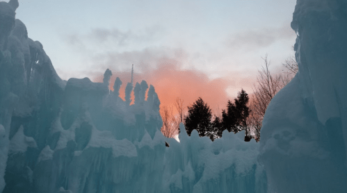 A winter scene with icy formations and a pinkish sunset sky peeking through trees.