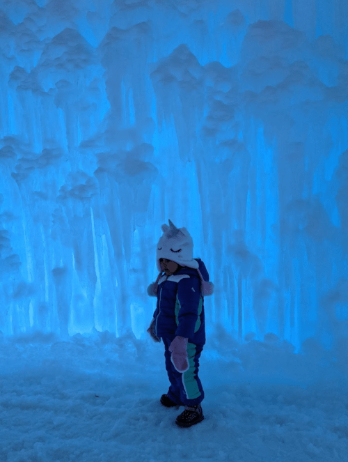 A child in a winter outfit stands near blue-lit ice formations, wearing a cute animal hat.