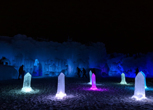 Colorful ice sculptures illuminated in blue and pink light, with silhouettes of people exploring the icy landscape at night.