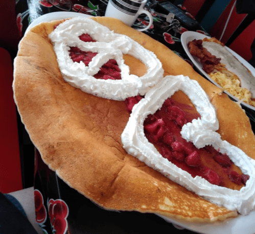 A large pancake shaped like two hearts, topped with raspberry filling and whipped cream, on a colorful tablecloth.