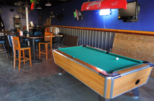 A pool table with a green felt surface in a casual bar setting, featuring wooden chairs and a dartboard on the wall.