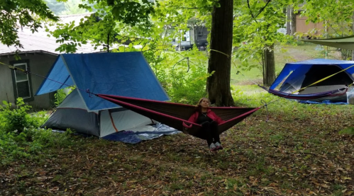 A person relaxes in a hammock between trees, with tents set up nearby in a wooded area.