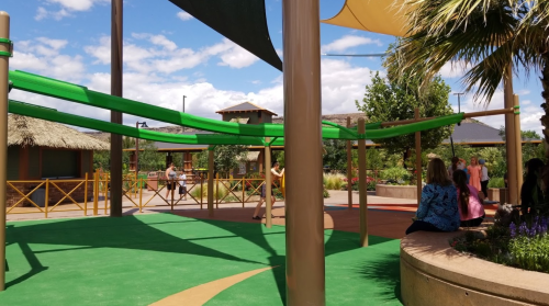 A colorful outdoor play area with green canopies, palm trees, and people enjoying the space under a blue sky.