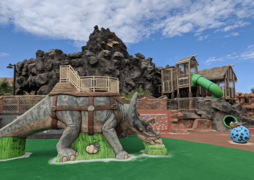 A dinosaur-themed playground featuring a triceratops sculpture, a climbing structure, and a green slide against a rocky backdrop.