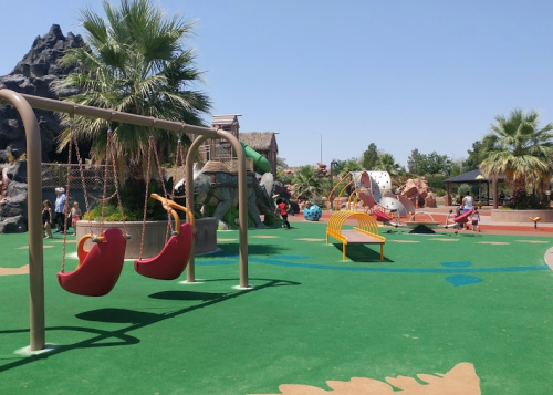 Colorful playground with swings, climbing structures, and palm trees under a clear blue sky. Children playing in the background.