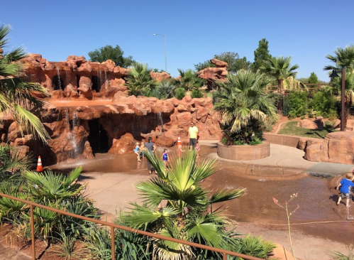Children play in a splash pad area with a rock formation and palm trees under a clear blue sky.