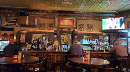 A cozy bar interior with patrons seated at the counter, shelves of liquor, and vintage decor. A TV displays a sports game.