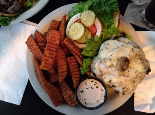 A plate with a cheeseburger topped with mushrooms, lettuce, tomato, pickles, and sweet potato fries on the side.