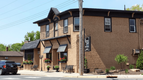 A two-story building with a textured exterior, flower boxes, and benches outside, set on a sunny street.