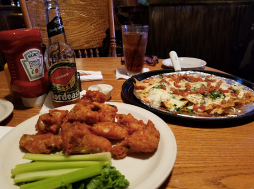 A plate of crispy chicken wings with celery, and a platter of nachos topped with cheese, tomatoes, and greens.