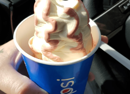 A hand holding a cup of soft serve ice cream with chocolate syrup on top, set against a car interior background.