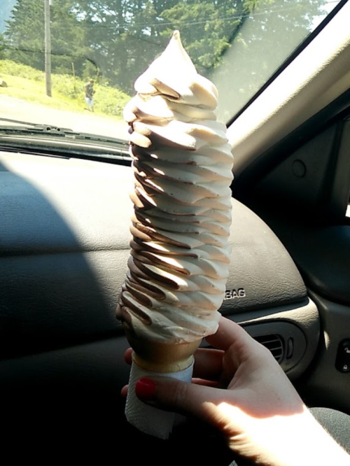 A hand holding a tall, swirled ice cream cone with chocolate and vanilla flavors inside a car.