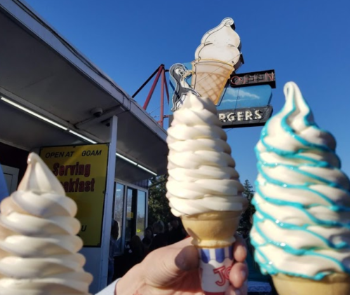 Three hands hold ice cream cones with swirled soft serve, in front of a retro ice cream shop sign.