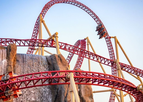 A roller coaster with red tracks loops over a waterfall against a clear sky.