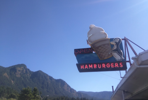 A vintage neon sign featuring an ice cream cone and the word "HAMBURGERS" against a clear blue sky and mountains.