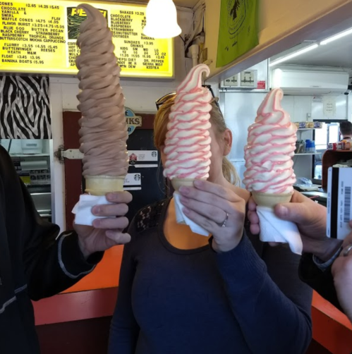 Three people hold up large ice cream cones in a colorful ice cream shop, showcasing chocolate and strawberry swirls.