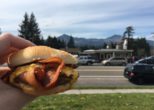 A hand holding a bacon cheeseburger in front of a scenic mountain backdrop and a roadside diner.