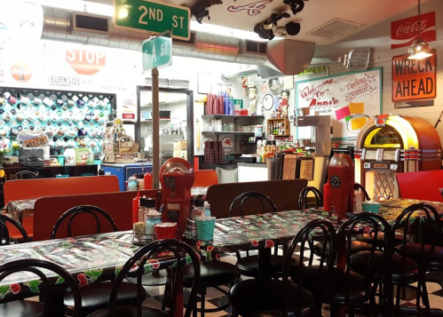 A retro diner interior with colorful decor, checkered floors, and a jukebox, featuring tables and vintage signage.