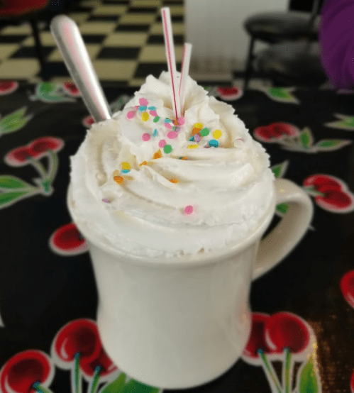 A mug filled with whipped cream, colorful sprinkles, and two straws, set on a cherry-patterned tablecloth.