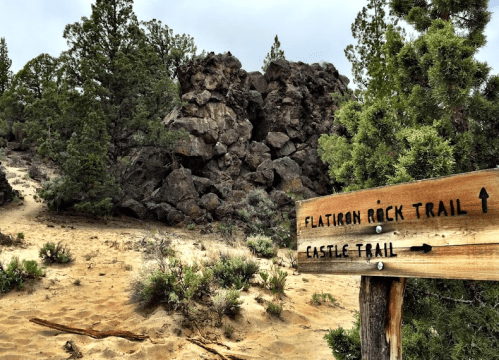 Wooden signpost indicating Flatiron Rock Trail and Castle Trail, with rocky terrain and pine trees in the background.