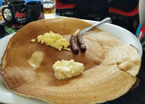 A large pancake on a plate with scrambled eggs, two sausage links, and a dollop of butter.