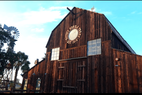 A rustic wooden barn with a circular sign, windmill, and palm trees under a clear blue sky.