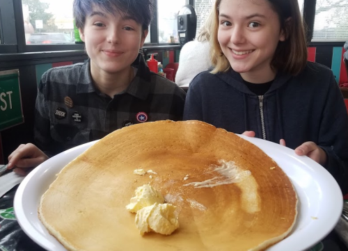 Two smiling friends hold a large pancake on a plate, with butter on top, in a cozy restaurant setting.