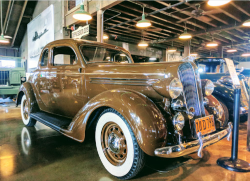 A vintage brown coupe with whitewall tires displayed in a museum setting.