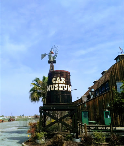 A rustic car museum sign on a large barrel, with a windmill and palm trees in the background under a blue sky.