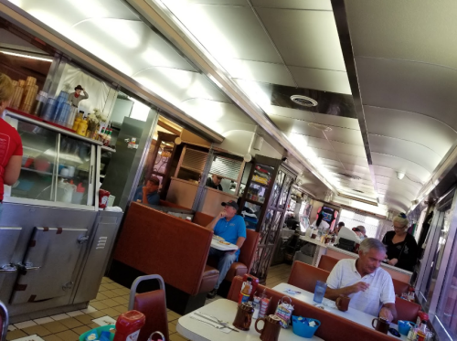 A retro diner interior with booths, tables, and patrons enjoying meals, featuring a counter and vintage decor.