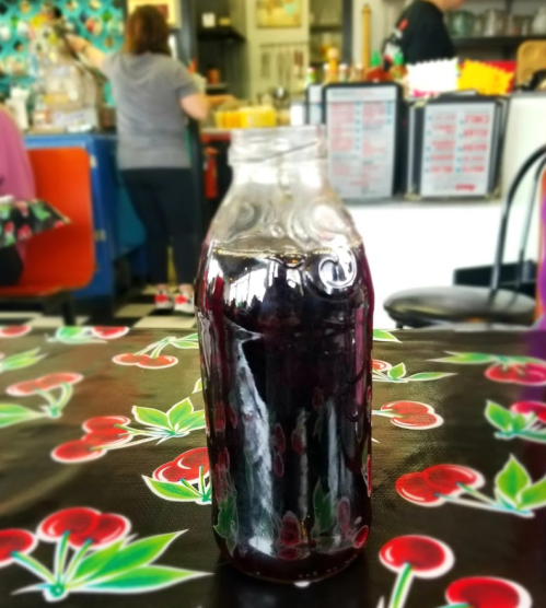 A glass bottle filled with dark liquid sits on a cherry-patterned tablecloth in a colorful café.