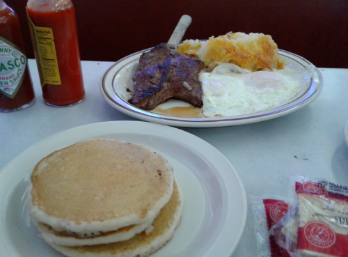 A plate of steak, eggs, and hash browns next to a stack of pancakes with icing on a table. Ketchup bottles in the background.