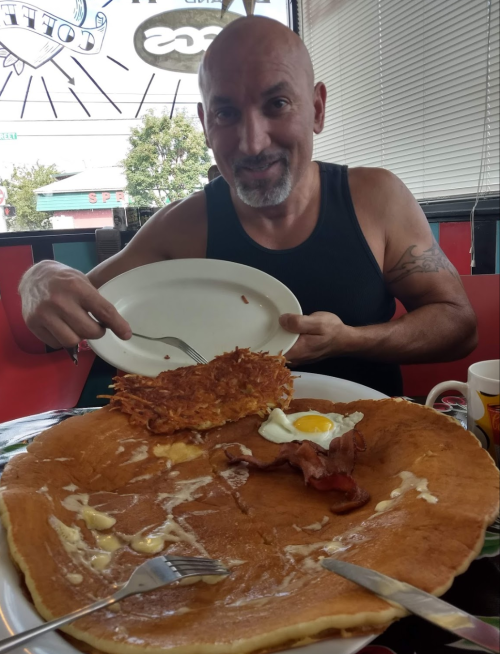 A man in a tank top smiles while holding a plate of large pancakes, bacon, an egg, and hash browns at a diner.