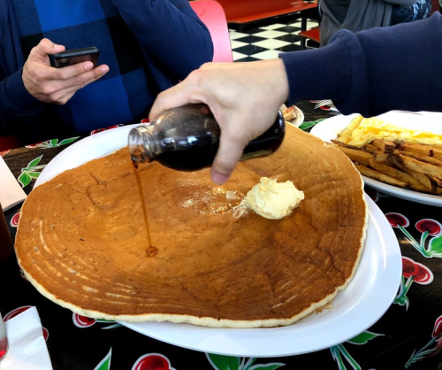 A large pancake on a plate with butter, as syrup is poured over it. A person is holding a phone in the background.