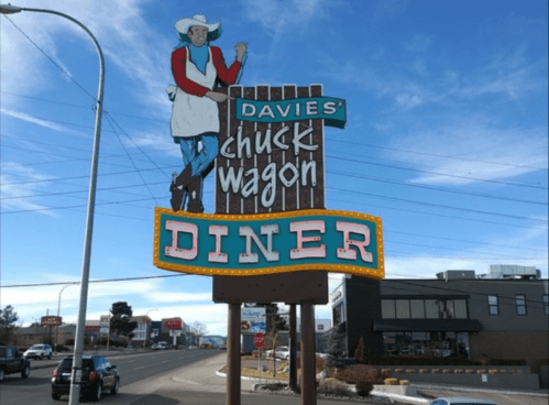 Sign for "Davies' Chuck Wagon Diner" featuring a cowboy figure, set against a blue sky and roadside scenery.