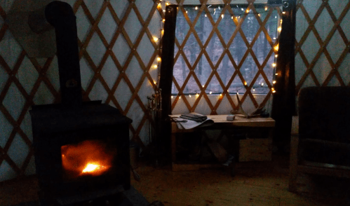 Cozy interior of a yurt with a wood stove, a table, and fairy lights illuminating a window.