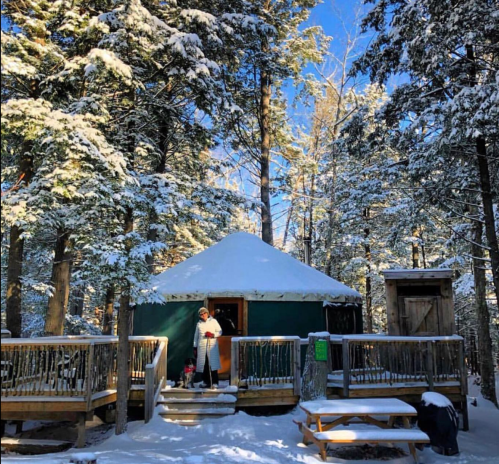 A cozy yurt surrounded by snow-covered trees, with a person standing on the deck in winter attire.