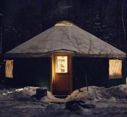 A cozy yurt illuminated at night, surrounded by snow and trees, with a warm glow from the door and windows.