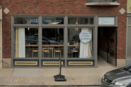Exterior of Happy Chicks Bakery, featuring large windows, a sign, and a small outdoor seating area.