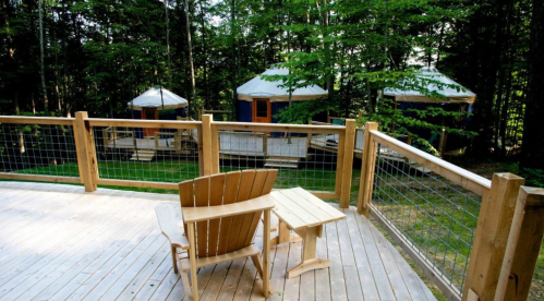 A wooden deck with a chair and table overlooks three yurts nestled in a forested area.